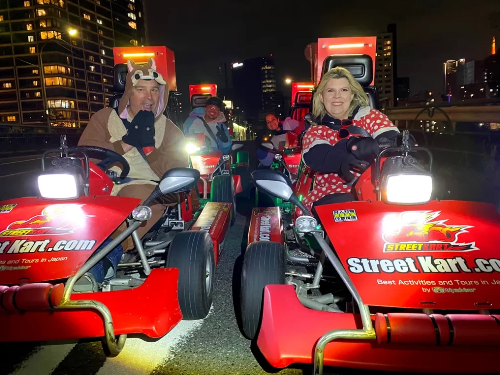 IMG_4263 - StreetKart Nighttime go-kart lineup on a city street; two costumed riders in front, one in a reindeer suit and the other in a red polka-dot jacket, smiling.