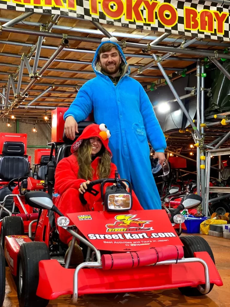 IMG_9935 - StreetKart Two people in red and blue Sesame Street onesies pose on a red go-kart indoors; sign reads Street Kart.